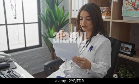 Junge hispanische Ärztin in einem Klinikbüro, in einem weißen Mantel gekleidet, sorgfältig medizinische Dokumente gelesen, während sie eine Brille hält, mit einer Stethosole Stockfoto