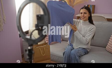 Frau zeigt blauen Pullover in gemütlichem Wohnzimmer mit Ringlicht, brünette Content Creator filmt Vlog zu Hause. Stockfoto