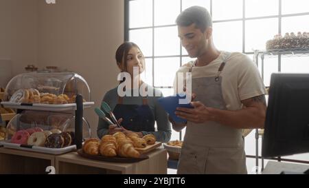 Mann und Frau in der Bäckerei, die Schürzen tragen und mit ausgestellten Backwaren zusammenarbeiten und in Innenräumen ein digitales Tablet verwenden Stockfoto