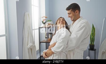 Mann und Frau in weißen Bademänteln lächeln und umarmen in einem Wellness-Spa-Raum mit hellem Tageslicht durch die Fenster und gemütlicher Inneneinrichtung Stockfoto