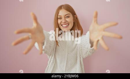 Fröhliche junge Frau mit rotem Haar in einem weißen Pullover, der vor rosafarbenem Hintergrund zur Kamera streckt. Stockfoto