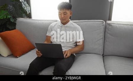 Junger Mann in seinem Wohnzimmer, der einen Laptop benutzt, während er auf einem bequemen Sofa sitzt, umgeben von moderner Wohnkultur und üppigen Pflanzen im Innenbereich, wodurch ein C Stockfoto