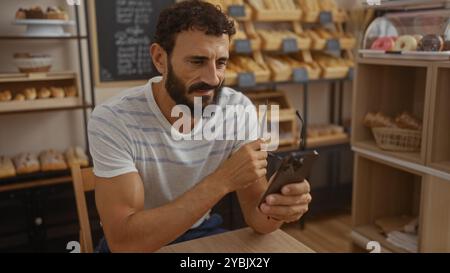 Hübscher Mann mit Bart in der Bäckerei, der auf das Telefon blickt und eine Brille hält Stockfoto