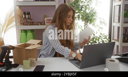 Junge Frau, die an einem Laptop in einem Wohnhaus arbeitet, umgeben von Paketen und Produkten, und die Dokumente am Schreibtisch sorgfältig durchsucht. Stockfoto