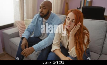 Interrassisches Paar, das drinnen in einem Wohnzimmer zusammen sitzt, Frau mit Hand auf Gesicht, beide schauten nachdenklich aus Stockfoto