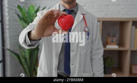 Ein hispanischer Mann in einer Klinik, der ein rotes Herz hält und einen Gesundheitsexperten darstellt, der die Herzgesundheit fördert. Stockfoto