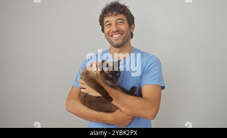 Ein fröhlicher junger hispanischer Mann in einem blauen Hemd, der eine siamesische Katze hält, isoliert vor weißem Hintergrund. Stockfoto