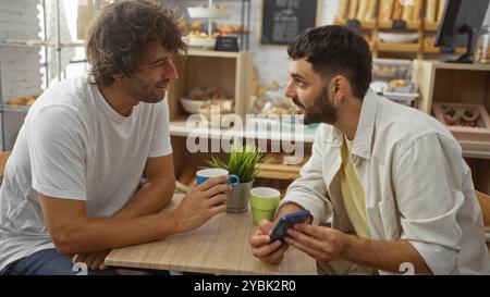 Männer, die in einer Bäckerei über Kaffee reden, über etwas auf einem Smartphone reden, während sie sich in einem Innencafé amüsieren Stockfoto