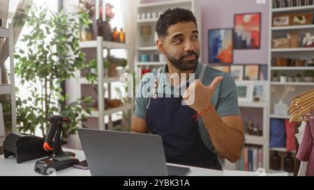 Junger hispanischer Mann in Schürze, der im Wohnkultur-Laden arbeitet, der auf Regale mit Pflanzen und Kunst im Hintergrund zeigt Stockfoto