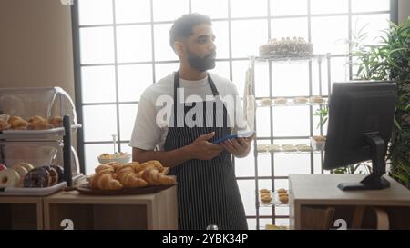 Junger hispanischer Mann, der in einer Bäckerei arbeitet und eine Tafel hält, während er neben einer Präsentation von Gebäck und Backwaren steht Stockfoto