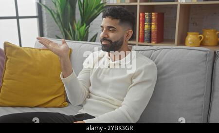 Hübscher hispanischer Mann mit Bart, der lässig auf dem Sofa im gemütlichen Wohnzimmer sitzt und lächelt Stockfoto
