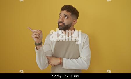 Hübscher hispanischer Mann mit Bart in der Schürze, der vor gelbem Hintergrund zur Seite zeigt Stockfoto