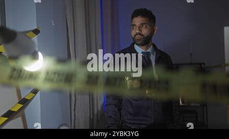 Ein ernsthafter hispanischer Mann mit Bart ermittelt einen Tatort in einem Haus, der mit gelben Polizeilinien in einem schwach beleuchteten Wohnzimmer aufgezeichnet wurde. Stockfoto