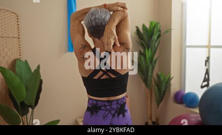 Reife Frau mit grauen Haaren, die sich im Turnsaal dehnt, in sportlicher Kleidung und mit Rückenmuskeln und Tätowierungen drinnen Stockfoto
