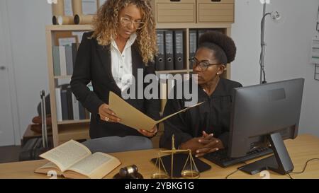 Frauen, die in einem Arbeitsbüro mit juristischen Dokumenten zusammenarbeiten, eine sitzt als Richter in einem schwarzen Gewand, während die andere neben ihrem Haft steht Stockfoto