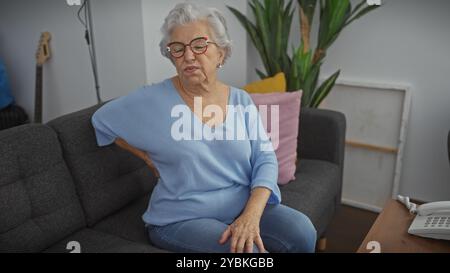 Ältere Frau mit Brille, die Rückenschmerzen hat, während sie zu Hause auf einer Couch sitzt. Stockfoto