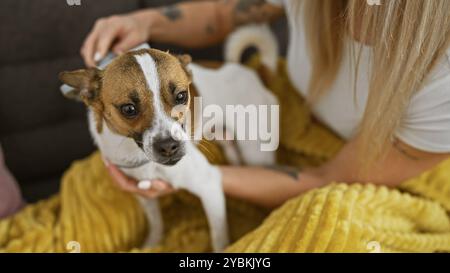 Eine blonde Frau kuschelt sich drinnen mit ihrem aufmerksamen Jack russell Terrier und liegt auf einer gemütlichen gelben Decke. Stockfoto