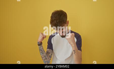 Ein hispanischer Mann mit Bart und Tätowierungen feiert siegreich vor gelbem Hintergrund Stockfoto