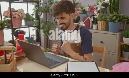 Ein fröhlicher junger Mann mit Bart feiert den Erfolg in einer lebendigen Blumenläden-Umgebung, die mit einem Laptop interagiert. Stockfoto