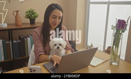 Hispanische Frau mit ihrem maltesischen Hund, der an einem Laptop in einem modernen Wohnzimmer arbeitet. Stockfoto