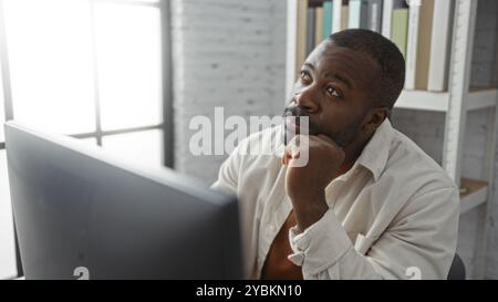 Junger Mann in einem Büro denkt tief nach, während er auf einen Computermonitor blickt, und schlägt Kontemplation und Konzentration in einem professionellen Innenbereich vor. Stockfoto