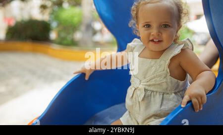 Niedliches blondes Kleinkind, das an einem sonnigen Tag auf einer blauen Rutsche auf einem Spielplatz im Park ihre Zeit genießt. Stockfoto