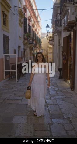 Eine junge hispanische Frau in einem weißen Kleid geht selbstbewusst an einem sonnigen Tag eine charmante Straße in gallipoli, apulien, italien hinunter. Stockfoto