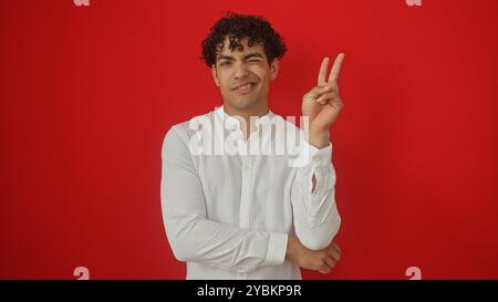 Junger hispanischer Mann, der zuzwinkernd und mit Gesten versehenes Friedenszeichen isoliert über rotem Hintergrund. Stockfoto