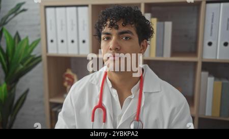 Ein selbstbewusster junger hispanischer Arzt mit lockigen Haaren, der ein Stethoskop trägt, posiert an seinem Arbeitsplatz und vermittelt einen Eindruck von Professionalität. Stockfoto