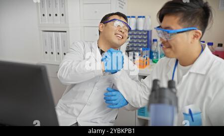 Zwei lächelnde Männer in Labormänteln und Schutzbrille, die in einem Labor einen freundlichen Faustschlag geben. Stockfoto