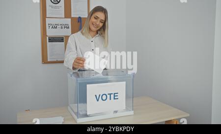 Eine junge, attraktive blonde Frau wählt in einem Wahlstudienzimmer und wirft ihren Stimmzettel in eine transparente Box mit einem Stimmschild während der Stockfoto