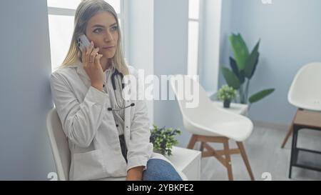 Eine junge Ärztin mit blonden Haaren in der Lobby der Klinik, die am Telefon spricht und ein Stethoskop um den Hals trägt und in der Sonne nachdenklich aussieht Stockfoto
