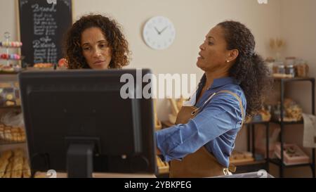Frauen arbeiten in einem Bäckereiraum zusammen, konzentrieren sich auf einen Computer, umgeben von Backwaren und bunten Macarons Stockfoto