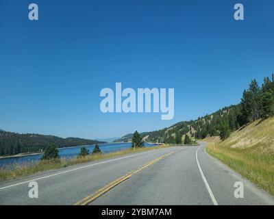 Eine malerische Straße führt entlang eines ruhigen Sees mit bewaldeten Hügeln unter einem klaren blauen Himmel. Stockfoto