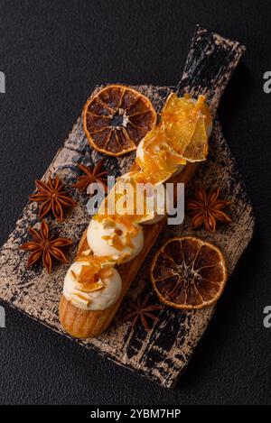 Süße Desserteclairs oder Profiterolen mit Schokoladenfüllung, bedeckt mit Sahne und getrockneten Orangen auf dunklem Betonhintergrund Stockfoto