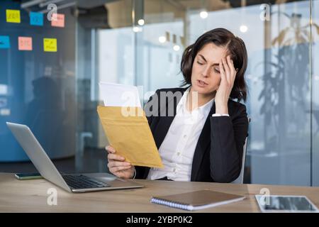 Gestresste Geschäftsfrau im Büro, die Dokumente liest und den Kopf hält. Der Ordner in der Hand deutet auf schlechte Nachrichten oder finanziellen Stress hin. Laptop und Notebook auf dem Schreibtisch in einem modernen Büro. Stockfoto