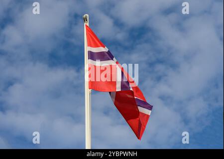 Die leuchtend rote norwegische Flagge flattert anmutig in der sanften Brise, vor dem Hintergrund eines klaren blauen Himmels mit weichen, weißen Wolken. Stockfoto