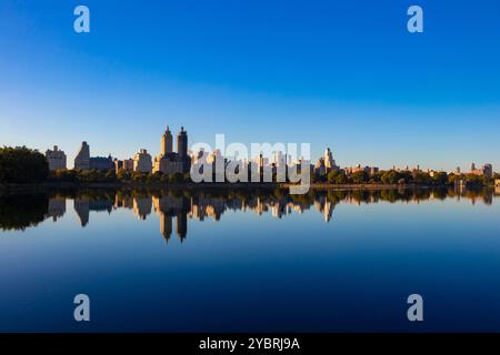 Die symettrische Skyline spiegelt sich während des Sonnenaufgangs im Central Park, New York, in ruhigem Wasser wider, wobei der klare blaue Himmel die Silhouette der Stadt unterstreicht Stockfoto