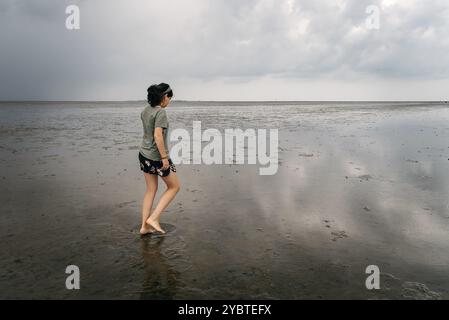 Junge Frau, die bei Ebbe allein an einem ruhigen und ruhigen Strand im Wattenmeer geht. Seitenansicht Stockfoto