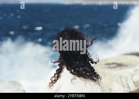 Junge Frau mit lockigen Haaren sitzt auf den Felsen und schaut auf das plätschernden Meer. Blick von hinten Stockfoto