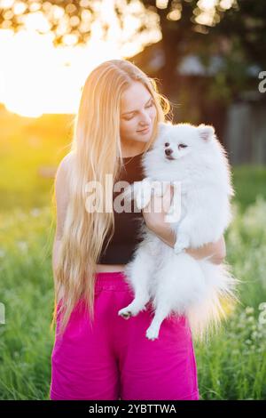 Die junge Frau umarmt ihren weißen pommerschen Hund in einem Feld bei Sonnenuntergang Stockfoto