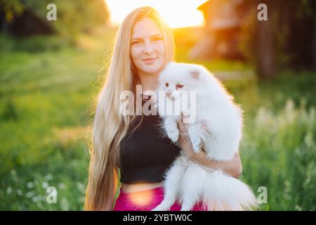 Die junge Dame umarmt ihren weißen pommerschen Hund auf einem Feld bei Sonnenuntergang Stockfoto