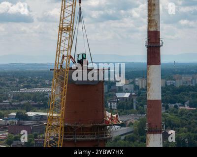 Arbeiten Sie oben auf dem Kamin. Ein hängender Bagger an einem Kran zerstört einen Schornstein. Drohnenansicht des Abbruchs des Schornsteins, Bagger arbeitet mit Rauch Stockfoto