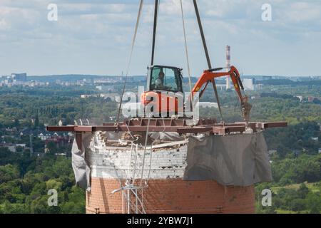 Arbeiten Sie oben auf dem Kamin. Ein hängender Bagger an einem Kran zerstört einen Schornstein. Drohnenansicht des Abbruchs des Schornsteins, Bagger arbeitet mit Rauch Stockfoto