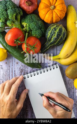 Frisches Gemüse, Brokkoli, Tomaten, Bananen, Kürbis, Zucchini, Kiwi und Zitrone, zwei Hände und ein Notizblock, Konzept für gesundes Essen und Neujahrsvorsatz Stockfoto