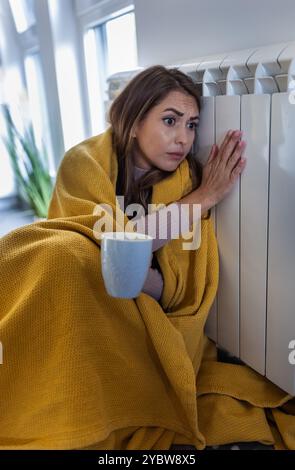 Hübsches Mädchen, das friert, auf dem Boden sitzt, in Decke gehüllt, sich auf den Heizkörper lehnt und eine Tasse Tee hält Stockfoto