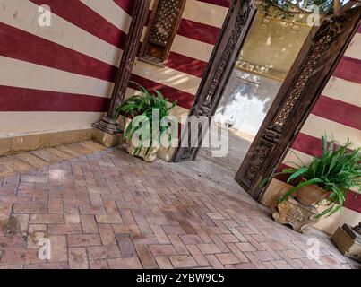 Ein Innenhof mit rot-weiß gestreiften Wänden, einem Backsteinboden und zwei Topfpflanzen. Eine Holztür führt zu einem anderen Innenhof Stockfoto