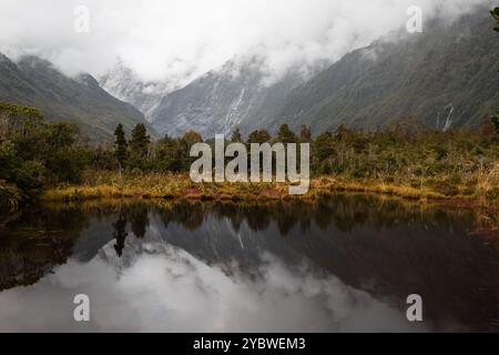 Neblige Reflexionen auf dem kleinen See mit schneebedecktem Franz Josef Gletscher im Hintergrund und große Wolken, die über Berge Rollen (Südinsel, New Zeala Stockfoto