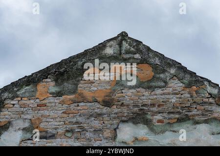 Abgenutzt und verwittert: Beschädigte Mauer und Dach eines alten Hauses gegen einen bewölkten Himmel. Stockfoto