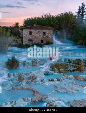 Besucher genießen das warme, mineralreiche Wasser der Saturnia Thermalbäder, umgeben von der atemberaubenden toskanischen Landschaft in der Abenddämmerung. Die leuchtend blauen Pools stehen im Kontrast Stockfoto